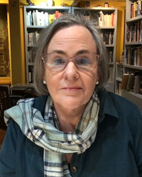 Catherine Ettinger, a woman with shoulder-length gray hair and glasses, wears a scarf and blue jacket, with books in the background