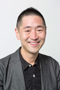 Ken Oshima, an Asian man with short black hair, wears a black shirt, gray blazer and smiles at the camera